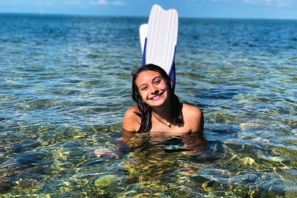 Photo of a teen girl in shallow water. She's on the ocean floor with her head and feet outside of the water, and she's wearing swimming fins. She's smiling as she faces the camera.